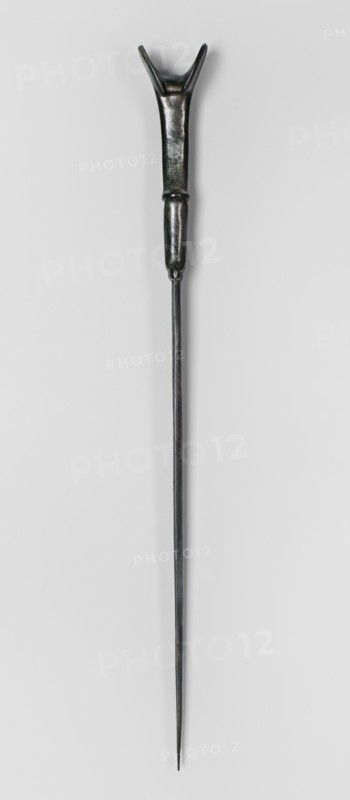 Dagger, unknown Luristan workshop. Dagger with a cast blade and a mushroom-shaped hilt with a rimmed mount; the raised edges of the mount served to hold a lost hilt casing, which may have been made of bone, wood, horn, or stone. The hilt shaft is richly profiled and flows directly into the blade with a centric point. Behind the short, rectangular base, blunted on both sides, the double-edged blade tapers evenly toward the tip. A slightly convex ridge runs through the center of the blade and splits along one-third of its length toward the base, creating a triangular flat field. 9th–6th century BC; Iron Period II 1st half of the 1st millennium BC. Luristan, Iran. Weapon. Height 33.8 cm; width 2.9 cm; depth 0.8 cm; weight 194 g., A long slender metal object with a dark, metallic finish that shows areas of black, dark gray, and hints of green; the top end has a forked, Y-shaped section attached to a thicker handle-like portion that tapers into a rounded connector before transitioning into ...