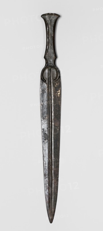 Dagger, unknown Luristan workshop. A dagger with a blade cast together with a mushroom-shaped hilt, with a rimmed mount whose raised edges served to hold a hilt casing that has not been preserved. The casing may have been made of bone, wood, horn, or stone. In the central part of the mount, short arched sheet-metal protrusions are bent inward to secure the casing. The hilt merges directly into a lanceolate, double-edged blade with a central point. The base of the blade is in the form of a convex ring, open at the bottom. A convex ridge with narrow grooves on the sides runs through the center of the blade from tip to base. 9th–6th century BC; Iron Period II end of the 2nd millennium–first half of the 1st millennium BC. Luristan, Iran. Weapon. Height 28.5 cm; width 2.9 cm; thickness 1.1 cm; weight 116 g., A long metallic object with a dark brownish-black patina, showing areas of lighter brown and muted gray where the surface is worn, is centered against a light gray background; it tapers...