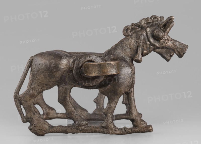 Bit, unknown Luristan workshop. The bit consists of a rigid bar and two cheek plates depicting a horse in profile. The bar, with a quadrangular cross-section, is slightly curved and has chamfered edges. Its ends are forged into a ribbon, which is wrapped in oppositely facing rings. The bar is inserted into two round holes in the cheek plates, which are located in the animals' torsos. Externally, they form circular rings protruding from the background, occupying the entire surface of the plates in this location. The cheek plates are modeled on one side; only the animal heads and necks are modeled on both sides. Attributed to Antoni Mierzejewski. 9th–6th century BC; Iron Period II c. 1000–601 BC. Iran, Luristan place of find and manufacture. Element of a horse harness. Height 8.6 cm; width 20 cm; depth 13.8 cm; weight 480 g., A small sculpted animal figure shown in profile on a flat surface, cast in a dark brown metallic material with a slightly rough, pitted texture and areas of lighter...
