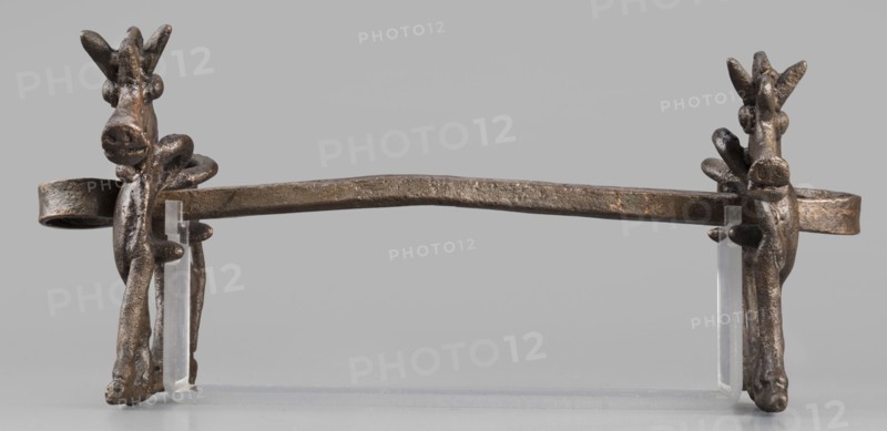 Bit, unknown Luristan workshop. The bit consists of a rigid bar and two cheek plates depicting a horse in profile. The bar, with a quadrangular cross-section, is slightly curved and has chamfered edges; its ends are forged into a ribbon wrapped in oppositely facing rings. The bar is inserted into two round holes in the cheek plates located in the animals' torsos; externally these form circular rings protruding from the background, occupying the entire surface of the plates at that location. The cheek plates are modeled on one side; only the animal heads and necks are modeled on both sides. Attributed to Antoni Mierzejewski, 9th–6th century BC; Iron Period II c. 1000–601 BC. Findplace/manufacture: Luristan, Iran. Element of a horse harness. Height 8.6 cm; width 20 cm; depth 13.8 cm; weight 480 g., A single metal horizontal bar with a textured, aged surface rests on a clear plastic support, flanked at each end by sculpted vertical elements that resemble stylized animal heads with elongat...