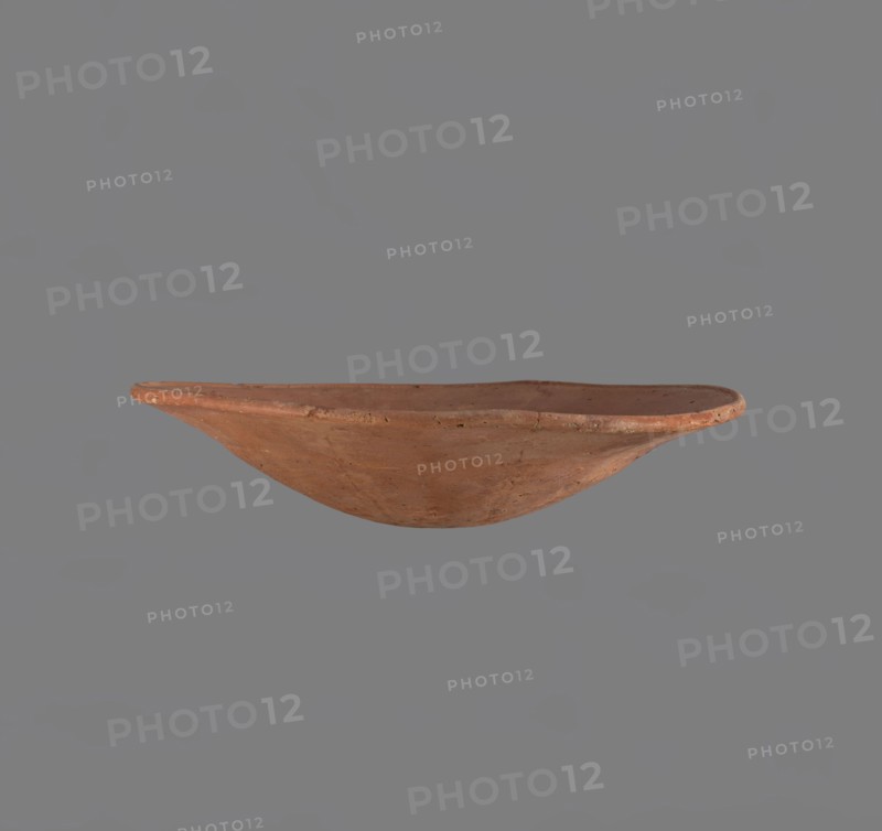 Bowl. Low bowl with irregular, strongly flaring walls and a rounded bottom. Rounded rim, slightly rolled outward. Surface smoothed and covered with a layer of red engobe. Bottom worked with a tool. Hand-made. Vessel glued together from several fragments. Egyptian pottery, clay vessel; ceramics, bowl. Comparative typology: Reisner, Mycerinus, type XXVII–XXVIII, fig. 72 2–5; c. 2686–2181 BC Old Kingdom, Edfu. Dimensions: height 6.1 cm; diameter rim 28.3 cm; diameter body 28.3 cm., A shallow, rounded clay bowl with an irregular rim is centered against a plain gray background; the bowl has a terracotta color with variations of reddish-brown and muted orange, showing a slightly rough texture and small surface imperfections.
