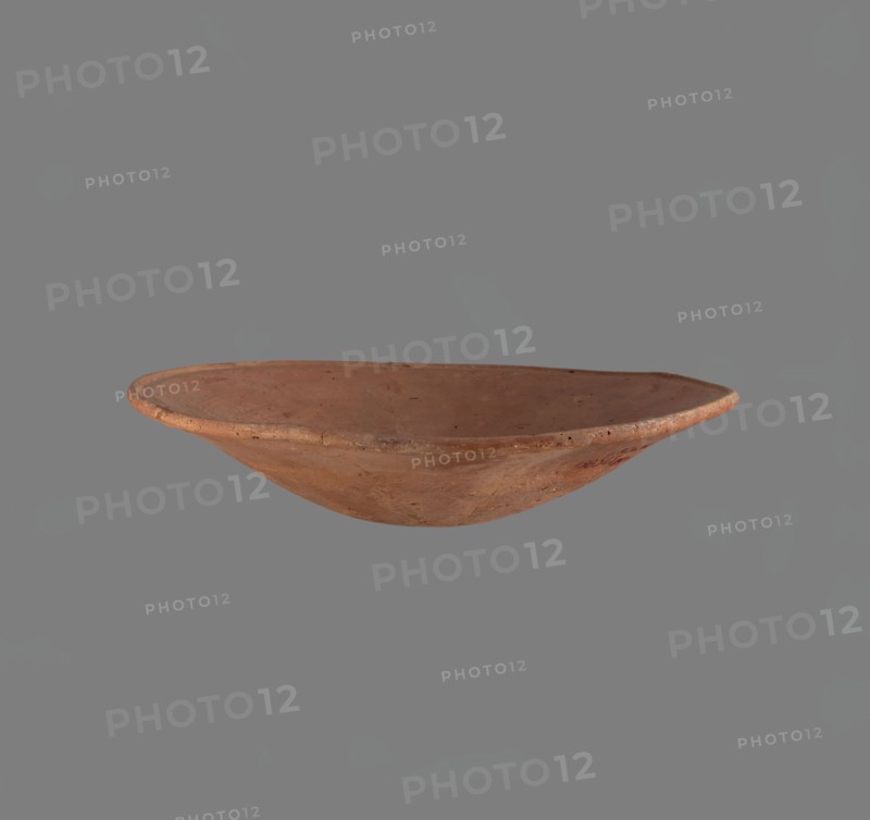 Bowl. Low bowl with irregular, strongly flaring walls and a rounded bottom. Rounded rim, slightly rolled outward. Surface smoothed and covered with a layer of red engobe. Bottom worked with a tool. Hand-made. Vessel glued together from several fragments. Egyptian pottery, clay/ceramic vessel, bowl. Old Kingdom, c. 2686–2181 BC. Height 6.1 cm; diameter rim/body 28.3 cm., Edfu place of origin., A shallow, round earthenware bowl with a slightly uneven rim and a smooth interior surface, shown against a plain gray background; the bowl appears in shades of reddish-brown and terra cotta with some darker speckles and subtle variations in tone, and the background is medium gray.