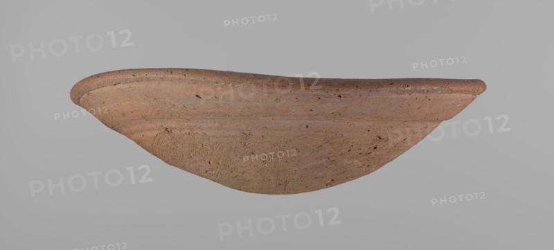 Shallow bowl of rounded, asymmetrical shape with a rounded, flaring, irregular rim. Surface is level; lower part finished to approximately half its height with a knife or similar tool. Slightly above the middle, a horizontal line likely from placement on a stand. Dark red clay; complete. From Edfu, local workshop. Old Kingdom, ca. 2686–2181 BC. Dimensions: height 7.2 cm, rim diameter 26.2 cm., A shallow, curved clay vessel displayed against a plain gray background, showing a rough, textured surface with visible tool marks and small pits; the vessel is primarily terracotta brown with variations including lighter beige and slightly darker brown areas, and there are hints of reddish and grayish tones in some spots.