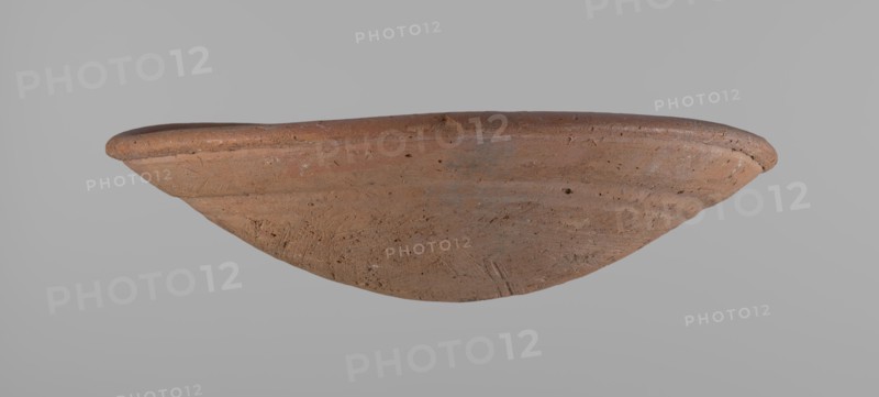 Shallow bowl of rounded, asymmetrical shape with rounded, flaring, irregular rim. Surface level; lower part finished to approximately half its height with a knife or similar tool. Slightly above the middle, a horizontal line likely from placement on a stand. Dark red clay; complete. From Edfu — local workshop. Comparative material: A. Reisner, A History of the City of the Giza Necropolis I, fig. 227, 35-8-16, ca. 2686–2181 BC Old Kingdom. Dimensions: height 7.2 cm, diameter 26.2 cm rim., A shallow, rounded ceramic bowl is shown from a side angle against a plain gray background; the bowl has an earthy brown color with variations of lighter and darker brown tones and small darker flecks, a slightly raised rim with a smoother texture, and a surface marked by subtle scratches, pits, and unevenness indicative of its material.