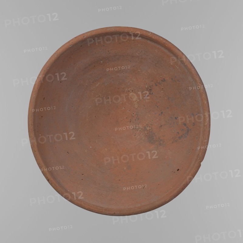 Bowl. Shallow, rounded, asymmetrical bowl with a rounded, flaring, irregular rim. Surface level; lower half of the interior finished with a knife or similar tool. Slight horizontal line slightly above mid-height likely from placement on a stand. Dark red clay; complete. From Edfu — local workshop. Comparative material: A. Reisner, Old Kingdom vessel ca. 2686–2181 BC, height 7.2 cm, rim diameter 26.2 cm., A round shallow bowl-like object fills the frame, showing a textured surface in shades of reddish-brown and terracotta with patches of darker brown and a few small black speckles; the rim is slightly raised and shows worn areas and tiny pits, and the background is a smooth light gray.