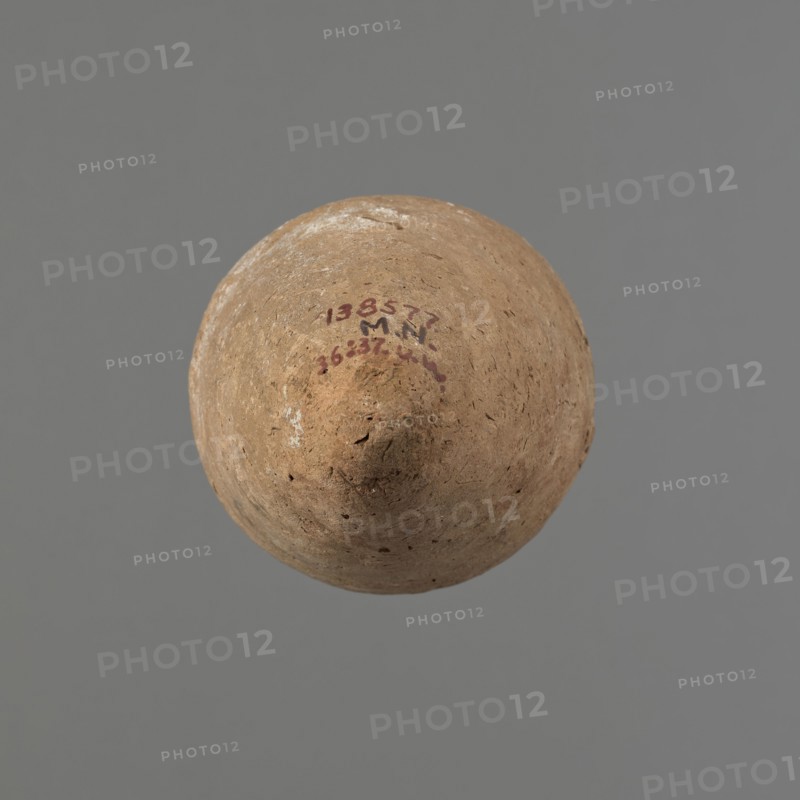 Vessel with hieratic inscription, Egyptian pottery. Conical clay vessel tapering toward the pointed bottom; irregular, widest at approx. 3/4 of the height. Narrow, sloping shoulders; short, wide neck; very gently rounded, slightly flaring rim. Handmade with the upper part neck and rim turned. Surface even, outer layer probably covered with engobe. Made of Nile clay NB2 Vienna System. In the central part of the belly are remains of a hieratic inscription in black probably sht dwdw. The vessel is complete and well preserved, with traces of salting on one side of the belly. Old Kingdom, ca. 2686–2181 BCE. Dimensions: height 16.1 cm; rim diameter 6.3 cm; body diameter 8.4 cm. Comparable examples: Petrie, Dendera; Hope, Akhmim type AIVa 2bi; Reisner; Brunton type 77N; Mace, Naga ed-Der; Petrie, Abydos II; Keiser, Avila, Dreyer, Jaritz, Laskowska-Kusztal, MDAIK 40., A rounded, slightly pointed object with a rough, weathered surface in shades of brown and tan is centered against a plain gray ...