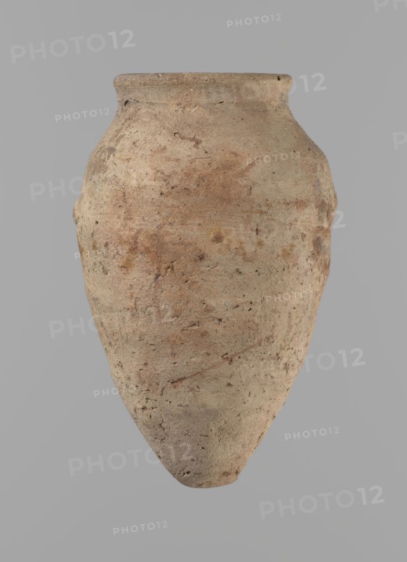 Vessel with hieratic inscription. Conical clay vessel tapering to a pointed bottom; irregular, widest at approx. 3/4 of the height. Narrow, slightly sloping shoulders; short, wide neck; very gently rounded, slightly flaring rim. Handcrafted; upper part shoulders, neck, rim turned. Smooth surface, externally probably covered with an engobe. Made of Nile clay NB2 Vienna System. Remains of a hieratic inscription in black on the lower part of the belly. Complete and well preserved. Comparative parallels cited; earlier dated to the 4th–5th Dynasty; Old Kingdom, ca. 2686–2181 BC. Dimensions: height 12.9 cm; rim dia. 5.5 cm; body dia. 7.8 cm., A roughly oval, tapered clay vessel with a narrow rounded rim, showing a weathered, textured surface in shades of beige, tan, brown, and muted reddish tones, set against a plain gray background; the vessel has a bulbous upper body that narrows to a pointed base and exhibits small pits, surface irregularities, and variations in color and roughness.