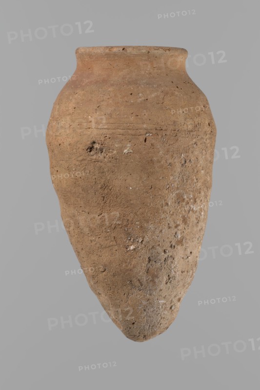 Vessel with hieratic inscription. Conical clay vessel tapering toward the bottom, irregular; widest at approx. 3/4 of the height. Narrow, only slightly sloping shoulders; short, wide neck; very gently rounded, slightly flaring rim; pointed bottom. Handcrafted; upper part shoulders, neck, rim turned. Smooth surface, exterior probably covered with engobe. In the widest part three shallow horizontal lines over approx. 0.8 cm. Made of Nile clay NB2 Vienna System. Remains of a hieratic inscription in black sht on the lower body. Vessel complete, well-preserved. Old Kingdom, ca. 2686–2181 BCE. Height 14.4 cm; rim diameter 5.4 cm; body diameter 8.4 cm. Comparative parallels cited to pottery from Dendera, Akhmim, Mycerinus-period contexts, Qau and Badari, Naga ed-Der, and Abydos., A single large clay vessel with an elongated tapered body and a rounded shoulder, shown against a plain gray background. The surface of the vessel is rough and pitted, with irregular texture and small inclusions visi...
