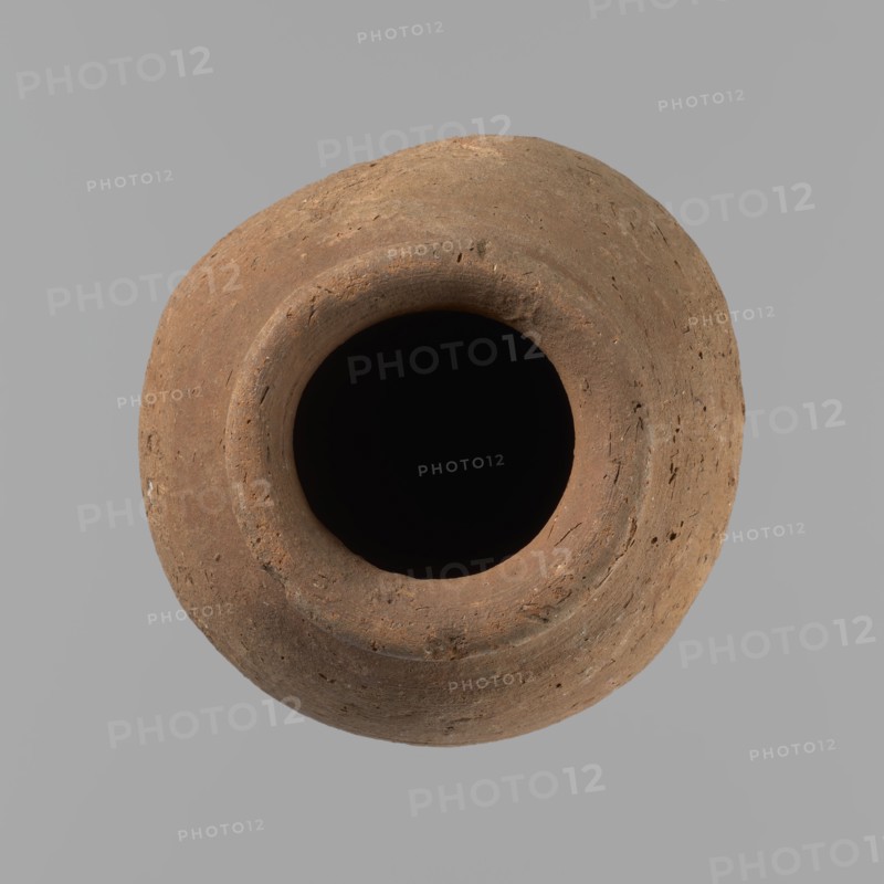 Vessel with hieratic inscription. Conical clay vessel tapering toward the bottom; irregular, widest at approx. 3/4 of the height. Narrow, only slightly sloping shoulders; short, wide neck; very gently rounded, slightly flaring rim; pointed bottom. Handcrafted: upper part shoulders, neck, rim turned. Smooth surface, externally probably covered with engobe. In the widest part three shallow horizontal lines placed over a space of approx. 0.8 cm. Made of Nile clay NB2 Vienna System. In the lower part of the body are remains of a hieratic inscription in black sht. Vessel complete, well-preserved. Comparative parallels cited; earlier dated to the Old Kingdom ca. 2686–ca. 2181 BC, find site Edfu. Dimensions: height 14.4 cm; rim diameter 5.4 cm; body diameter 8.4 cm., A top-down view of a round, open-mouthed clay vessel with a rough, textured surface in shades of brown and tan set against a uniform light gray background, showing the deep dark interior and the uneven rim with small pits and var...