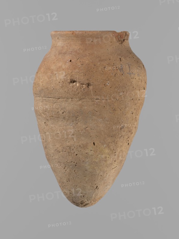 Vessel with hieratic inscription. Conical clay vessel tapering toward the bottom, irregular, widest at approx. 3/4 of the height. Narrow, only slightly sloping shoulders; short, wide neck; very gently rounded, slightly flaring rim; pointed bottom. Handcrafted with the upper part shoulders, neck, rim turned. Smooth surface, externally probably covered with an engobe. Made of Nile clay NB2 Vienna System. Remains of a hieratic inscription in black oak on the lower part of the belly. Complete and well preserved. Old Kingdom, ca. 2686–2181 BC; height 12.5 cm; rim diameter 5.4 cm; belly diameter 8 cm. Comparative parallels cited Petrie, Dendera; Hope, Akhmim; Reisner; Brunton; Mace; Petrie, Abydos; Keiser et al.., A single large earthenware vessel with a rounded shoulder tapering to a pointed base is centered against a plain gray background, showing a rough textured surface with small pits and abrasions; the vessel is primarily light brown with patches of darker brown and faint areas of yell...