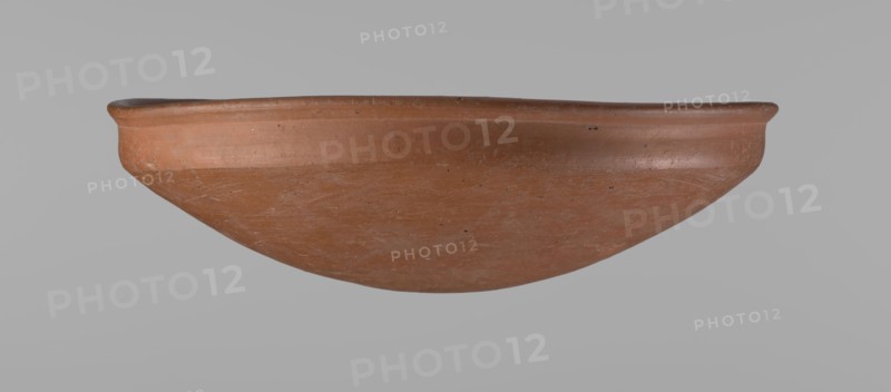 Medum-type bowl. Medum-type bowl with characteristic carination under the rim. Rounded bottom. Straight rim, slightly flaring. Narrow, very sloping shoulders — almost vertical. Diameter of the rim is larger than the diameter of the shoulders. Clay probably NB. Surface leveled and covered with a layer of engobe, then polished. Vessel color: red. Vessel complete, very well preserved. Old Kingdom late Old Kingdom, c. 2686–2181 BC. Edfu place of origin. Vessel height 7.5 cm; diameter 25.8 cm rim; diameter 25.2 cm shoulders. Comparative reference: Junker, Giza I, pl. I,2,16., A shallow rounded vessel with a smooth rim and curved body in terracotta tones of reddish-brown and orange, showing surface irregularities and fine scratch marks, with small darker speckles and subtle lighter areas, photographed against a plain light gray background.