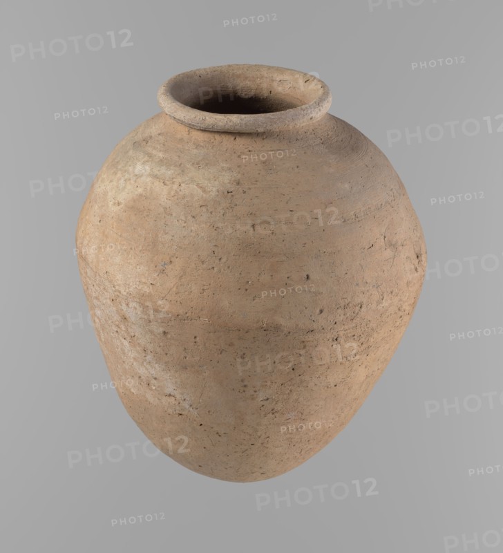 Vessel, Egyptian ceramics, clay vessel of ovoid, slightly downward-stretched shape, widest at about two-thirds of the height. Broad shoulders, very short wide neck; roller-shaped rim, oval in cross-section, slightly flaring; rounded bottom. Clay with significant organic admixture Viennese System. Handcrafted from two parts joined at about two-thirds height; upper part turned. Surface leveled. Traces of a white substance on outer surface and rim. Beige color. Complete, well-preserved. Late Old Kingdom, ca. 2686–2181 BC. From Edfu. Height 24.9 cm; rim diameter 10.2 cm; body diameter 20.7 cm., A single large rounded clay vessel with a wide rim and open mouth sits against a plain background, showing a textured surface with visible imperfections and tool marks; its color palette includes shades of light brown, tan, beige, and small areas of darker brown and gray, and the rim appears slightly lighter than the body.