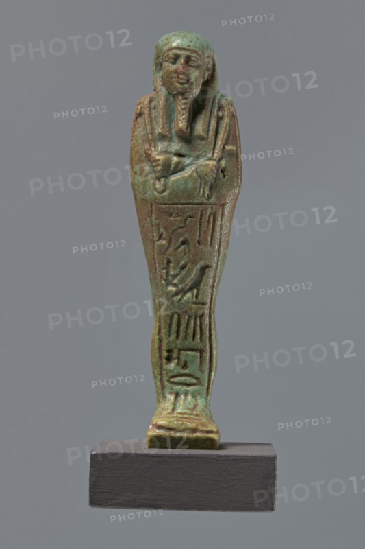 Ushabti Hora, Mummy-shaped figurine shabti, mummy-shaped figurine, standing on a base, back supported by a pillar/pilaster. Arms crossed on the chest, right folded over the left. Hoes in both hands. On the back is a basket, slung on a single string over the left shoulder. On the head is a three-part, smooth wig. Long braided beard. Face with proportional features, mouth set in a slight smile. Inscribed inscription on the front of the legs in one wide column framed with lines: Shd Osiris Hor born of Her-ib, justified by the voice. Ptolemaic Period, Egypt, 332–30 BC. Height 11.1 cm., A small statuette of a standing mummiform figure is shown against a plain grey background, displayed on a dark rectangular base; the figure has a stylized human face with a headdress and a false beard, arms crossed over the chest, and a narrow elongated body with carved relief details along the front. The surface shows a weathered greenish-blue patina with areas of brown and darker green, and a few spots of ...
