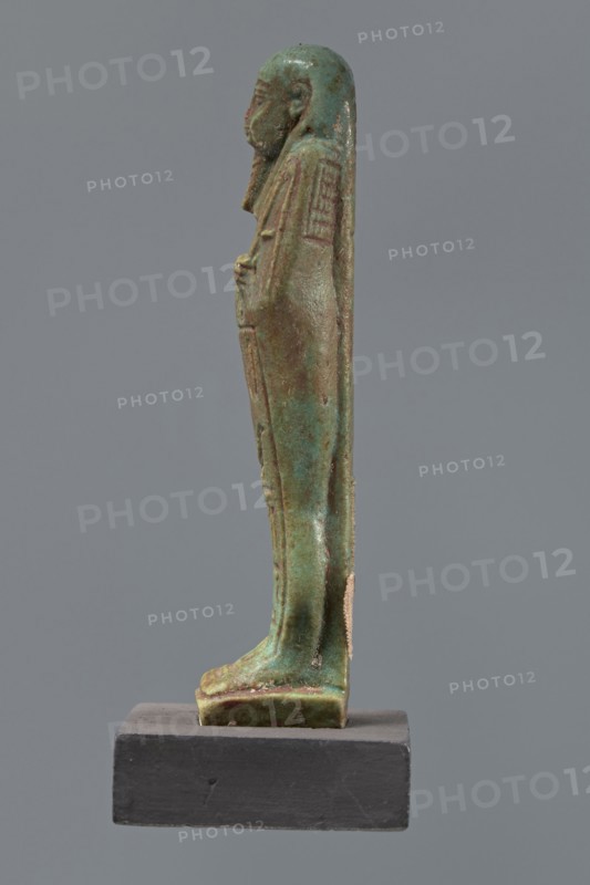 Ushabti Hora, Mummy-shaped figurine, funerary shabti, Ptolemaic Period, 332–30 BC, Egypt. Standing on a base, back supported by a pillar. Arms crossed on the chest, right over left, holding hoes in both hands. Basket on the back, slung on a single string over the left shoulder. Wearing a three-part smooth wig and a long braided beard. Face with proportional features and a slight smile. Inscribed on the front of the legs in one wide column framed with lines: Shd Osiris Hor born of Her-ib, justified by the voice. Height 11.1 cm., A slender statuette of a standing human figure displayed in profile on a dark rectangular base, with a smooth rounded head and elongated body draped in a long garment that reaches the feet; the surface shows a weathered, mottled appearance in shades of green and turquoise with patches of brown and darker green, and traces of a lighter beige material adhered to one side; the base is matte dark gray.