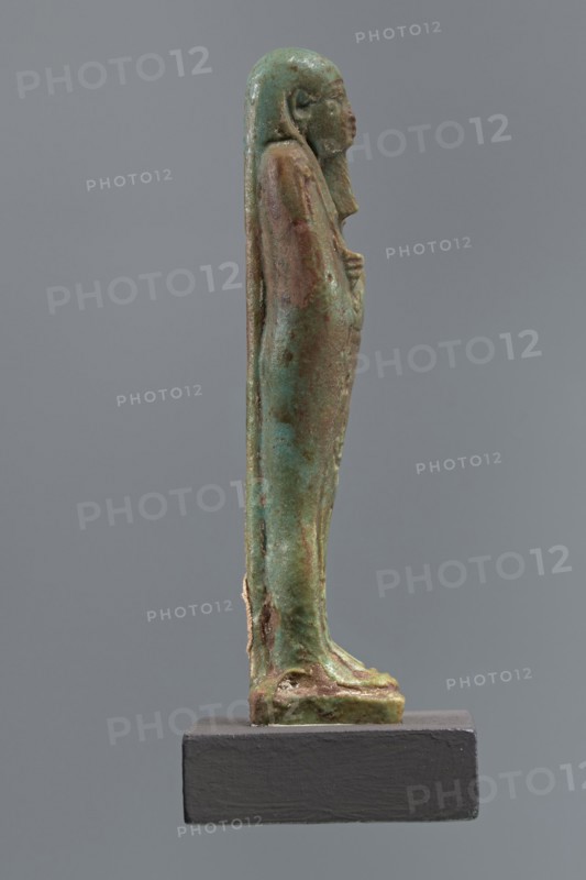 Mummy-shaped shabti of Horus inscribed Shd Osiris Hor born of Her-ib, justified by the voice, Ptolemaic Period, Egypt, 332–30 BC. Standing on a base with back supported by a pillar; arms crossed on the chest right over left holding hoes in both hands; a basket on the back slung over the left shoulder by a single string. Wears a three-part smooth wig and a long braided beard. Face with proportional features and a slight smile. Inscription in one wide column framed with lines on the front of the legs. Height 11.1 cm., A slender standing human figure sculpture shown in profile on a rectangular base, with a smooth elongated form and a head with discernible facial features and long hair or headdress flowing down the back; the surface shows varied green and brown patina with areas of lighter turquoise and darker verdigris, and some rougher brownish spots and worn textures, while the base is a solid dark gray.
