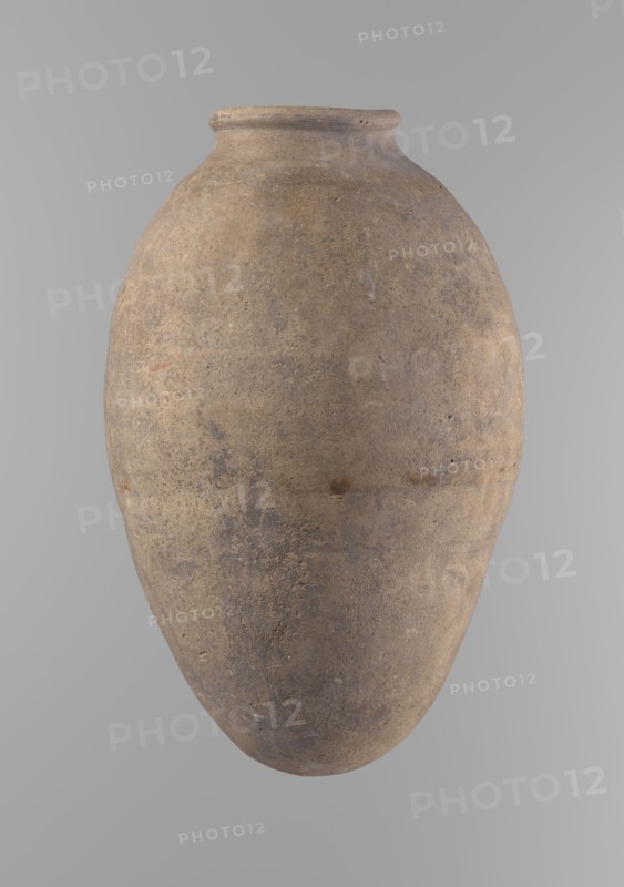 Vessel with an ovoid belly, widest about halfway up. Wide, short, almost cylindrical neck with a rounded, slightly tapering rim and rounded bottom. Made of Nile clay in two parts, joined about one-third of the vessel's height. Even surface, beige color, complete. Ceramic Egyptian vessel, clay vessel; height 35.5 cm, rim diameter 10.2 cm, body diameter 22.2 cm. Old Kingdom, Edfu, ca. 2686–2181 BC. Comparative typology: Reisner, Mycerinus, type V–XI,7 and XV–XVI., A large rounded ceramic jar with a narrow, slightly flared neck and a rounded base, shown against a plain light gray background; the jar surface appears textured and weathered with a mix of earthy browns, muted tans, and subtle grayish tones, with a few darker spots and faint variations in color across its surface.