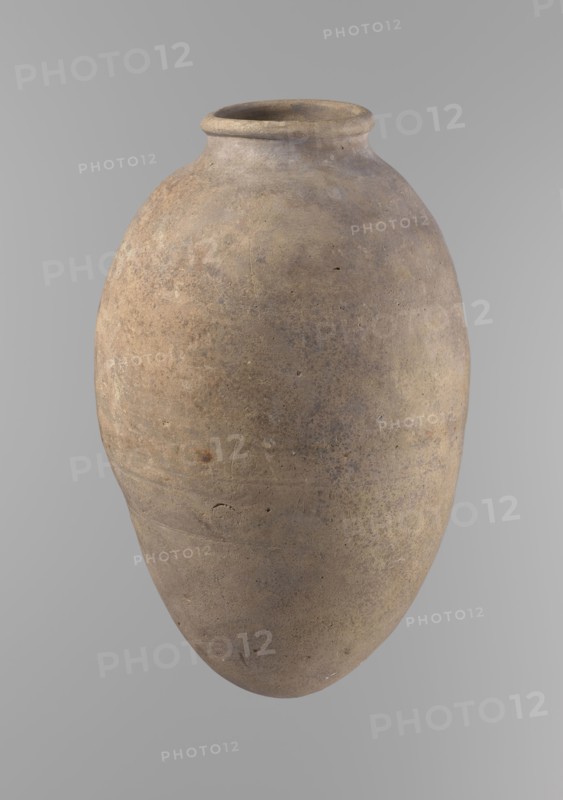 Vessel with an ovoid belly, widest about halfway up. Wide, short, almost cylindrical neck. Rounded, slightly tapering rim. Rounded bottom. Made of Nile clay in two parts joined about one-third of the vessel's height. Surface even, beige color. Complete. Ceramic Egyptian vessel, clay vessel; Old Kingdom, ca. 2686–2181 BC, Edfu. Height 35.5 cm; diameter 10.2 cm rim, 22.2 cm body. Comparative type: Reisner, Mycerinus, type V–XI,7 and XV–XVI., A large rounded clay vessel with a narrow neck and flared rim, shown against a plain light gray background; the vessel surface is rough and weathered with a mottled palette of brown, tan, beige, and hints of gray, displaying irregular patches, small pits, and subtle textured variations across its curved body.