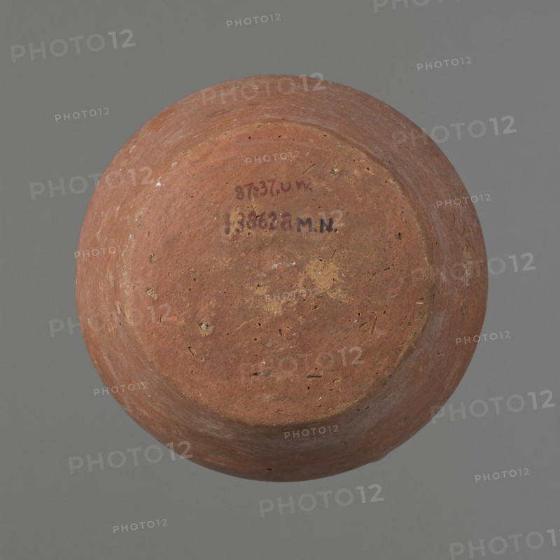 Jug, ceramics, Egyptian clay vessel jug — small jug with one handle; widest about 2/3 of the vessel's height. Wide, rather sloping shoulders; short neck widening toward the spout; straight, slightly flaring spout. Lower handle attached slightly above the vessel's widest diameter; upper attachment reaches the spout, protruding 1 cm above the spout line. Handle circular in cross-section on the lower part, slightly flattened in the upper part. Slightly convex bottom. Made of Nile clay. Surface even and covered with a thick layer of red slip on the outside and on the rim and neck inside. Color red-brown. Complete and fairly well preserved; slip worn away in places. Dating and origin: ca. 2686–2181 BC, Old Kingdom; Edfu. Measurements: height 12.7 cm; rim diameter 8.4 cm; body diameter 13.5 cm; bottom diameter 9.5 cm. Analogies: A. Fakhry, The Monuments of Sneferu at Dahshur, figs. 4–4., A round, shallow vessel viewed from above with a textured, worn surface showing earthy tones of reddish-b...