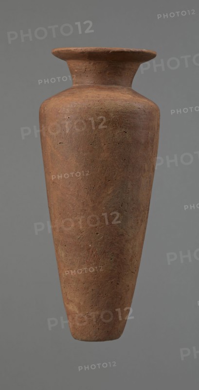 Vessel, Egyptian pottery, Type HS vessel, conical slender shape, widest at about 3/4 of the height. Shoulders wide, almost horizontal, slightly sloping. Neck wide, short, funnel-shaped in lower part, widening towards the rim from about half its height. Rim wide, flat on top, slightly rounded at bottom, slightly asymmetrical. Bottom flat, slightly asymmetrical. Neck made separately and glued to the shoulders. Surface even; lower part finished to approx. 3/4 of height with a knife or similar tool. Outer surface and inner surface of rim and neck covered with dark red engobe and polished. Color dark red. Reconstructed from many original fragments; gaps filled with plaster. c. 2686–c. 2181 BC Old Kingdom, Edfu. Vessel; height 24.8 cm, diameter rim 8.9 cm, diameter shoulders 10.3 cm, diameter bottom 4.1 cm., A tall, narrow vase with a flared rim and a tapered body suspended against a plain background; the surface shows a rough, textured finish with visible small pits and irregularities. The ...