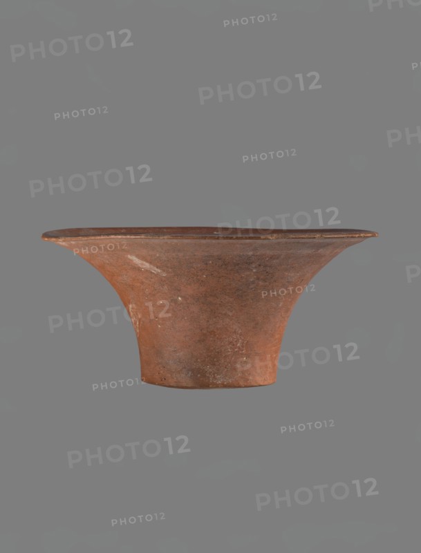 Bowl, Egyptian pottery, clay vessel — deep, cup-shaped bowl with a slightly convex bottom and sides arching upwards; straight rim, flat on top, flaring. Made of Nile clay NB1? and covered with an engobe containing red ochre and thoroughly polished. Vessel color: red. Well-preserved with slight damage to clay on the rim. Comparative material cites parallels from IV–VI dynasty and late VI dynasty contexts; typologically dated to the Old Kingdom ca. 2686–2181 BC. Place of origin: Edfu. Dimensions: height 13.8 cm; rim diameter 28.8 cm; bottom diameter 11.9 cm., A single wide-brimmed pottery bowl with a flared rim and a narrow base suspended against a neutral background; the vessel has an earthy reddish-brown color with patches of darker brown and small pale spots, a slightly rough textured surface showing subtle irregularities and wear, and the rim appears thinner and slightly darker than the body.