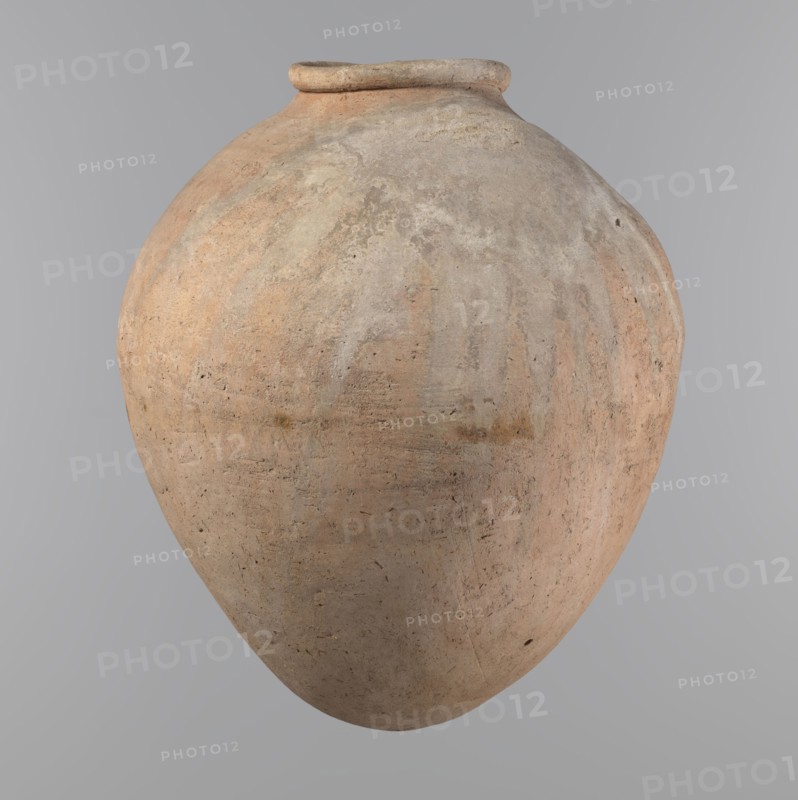 Vessel, Egyptian clay vessel of ovoid, slightly downward-stretched shape, widest at about 2/3 of the height; broad sloping shoulders, very short wide neck; roller-shaped rim oval in cross-section, slightly flaring; rounded bottom. Hand-made from two parts joined at about 2/3 of the height, upper part turned. Surface leveled, possibly covered with a layer of engobe; traces of a white substance in places on the outer surface and on the rim. Clay corresponds to NB2 Viennese system with a significant organic admixture. Colour beige. Well-preserved. Old Kingdom, ca. 2686–ca. 2181 BC; provenance Edfu. Vessel; height 34.6 cm, rim diameter 11.6 cm, body diameter 29 cm., A large rounded ceramic vessel with a narrow rim, showing a weathered, textured surface in shades of beige, tan, light brown, pale gray, and faint peach, with irregular patches and streaks of slightly darker and lighter tones and small surface pits and scratches against a plain neutral gray background.