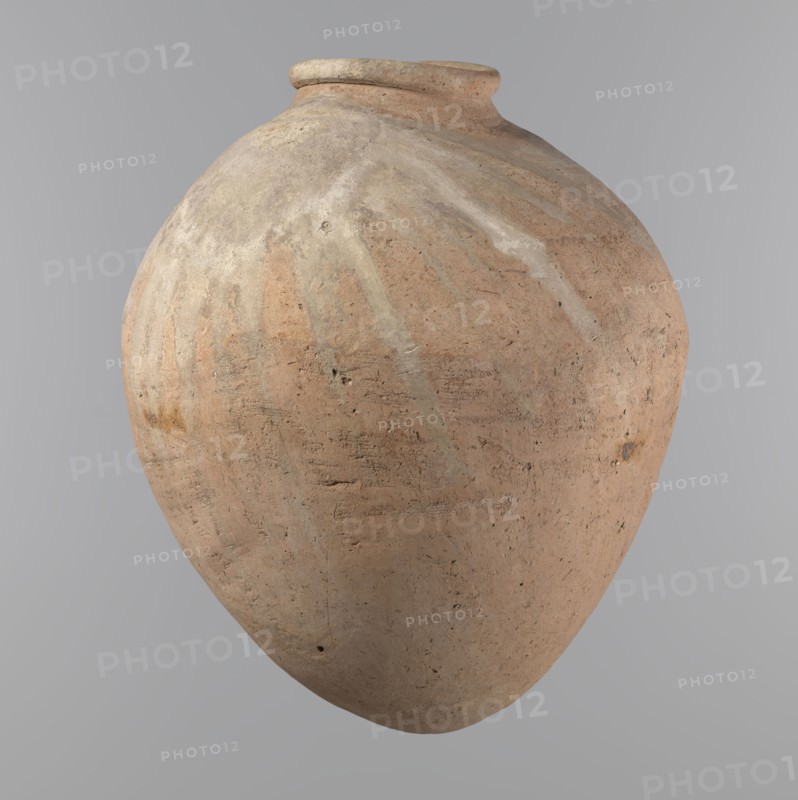 Vessel, Egyptian pottery vessel of ovoid, slightly downward-stretched shape, widest at about 2/3 of the height. Broad, sloping shoulders, very short wide neck. Roller-shaped rim, oval in cross-section, slightly flaring. Rounded bottom. Made of NB2 clay Viennese System with significant organic admixture. Hand-made from two parts joined at about 2/3 of the height, turned in the upper part. Surface leveled possibly covered with a layer of engobe. Traces of a white substance in places on the outer surface of the body and on the rim. Beige color. Well-preserved. Ca. 2686–2181 BC Old Kingdom, Edfu. Vessel; height 34.6 cm, rim diameter 11.6 cm, body diameter 29 cm., A large rounded clay vessel with a narrow rim and bulbous body, showing a textured surface with patches of light tan, beige, pale gray, and muted reddish-brown, faint streaks of lighter color radiating from the top, small pits and rough marks across its surface, and a neutral gray background.