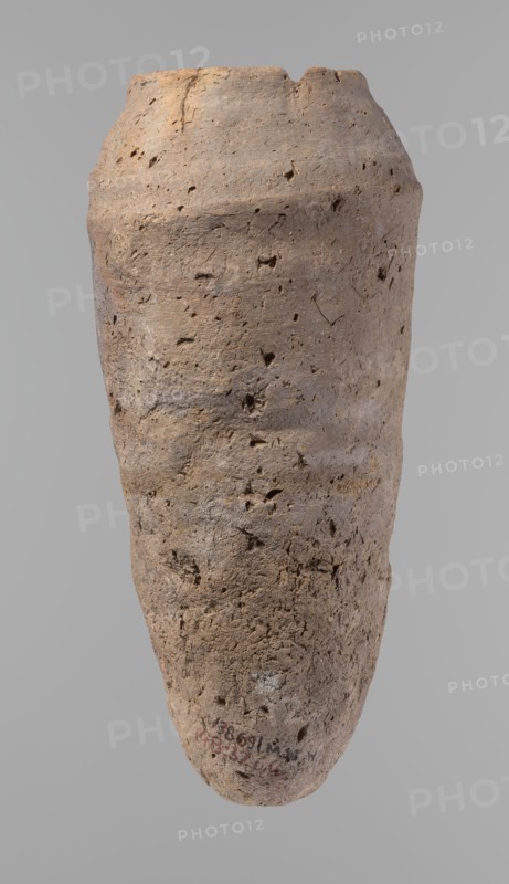 Vessel, Egyptian pottery, hand-made clay vessel; Old Kingdom, c. 2686–c. 2181 BC; from Edfu; vessel, height 33.5 cm, rim diameter 10.4 cm, body diameter 15.1 cm., A tall, weathered clay vessel with a rough, pitted surface occupies the image, showing an elongated, slightly tapered form with an irregular rim that has a small chip. The surface displays a range of earthy colors including light brown, tan, beige, and touches of gray, with darker brown spots and small shadowed cavities across the body. The texture appears coarse and porous with visible pits, cracks, and tiny holes, and there are subtle variations in hue creating faint horizontal banding and mottling. The background is a uniform neutral gray.