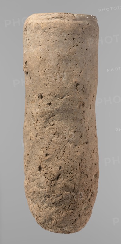 Vessel, Egyptian pottery, ceramics; almost cylindrical hand-made clay vessel, widest just below the rim, rounded bottom, slightly marked narrow sloping shoulders, straight tapering rim. Made of Nile clay with numerous organic inclusions and limestone particles; surface uneven, better worked in upper part, with numerous uneven depressions especially at the bottom. Brown color. Fairly well preserved; surface cracks attributed to careless kneading and firing. Old Kingdom, ca. 2686–2181 BC; from Edfu. Function: vessel. Height 36.5 cm; diameter 11.5 cm rim, 13.2 cm body., A tall cylindrical object with a rough, pitted surface standing against a plain light gray background; the object appears textured and uneven with a weathered, earthy surface showing small holes, indentations, and irregularities; its color is predominantly light brown with areas of darker brown and subtle tan variations; the top edge is slightly rounded and shows a few lighter spots, while the lower portion tapers slightly...