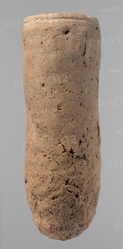 Vessel, Egyptian pottery, ceramics, clay vessel, hand-made. An almost cylindrical vessel, widest in its upper part just below the rim. The bottom is rounded. The shoulders are slightly marked, irregular, very narrow, and sloping. The rim is straight, tapering. Handmade of Nile clay with numerous organic inclusions and limestone particles visible on the surface. The surface is uneven and better worked in the upper part. Carelessly made, with numerous uneven depressions on the surface, especially at the bottom. Color: brown. Fairly well preserved; surface cracks attributable to careless kneading of the clay and firing. ca. 2686–ca. 2181 BC; Old Kingdom, Edfu. Vessel; height 36.5 cm; diameter 11.5 cm rim, 13.2 cm body., A tall, cylindrical object with a rough, porous surface in shades of light brown and tan, showing irregular pits, small indentations, and scattered darker brown spots; the surface texture varies from smoother areas near the top to more cracked and crumbly regions toward th...