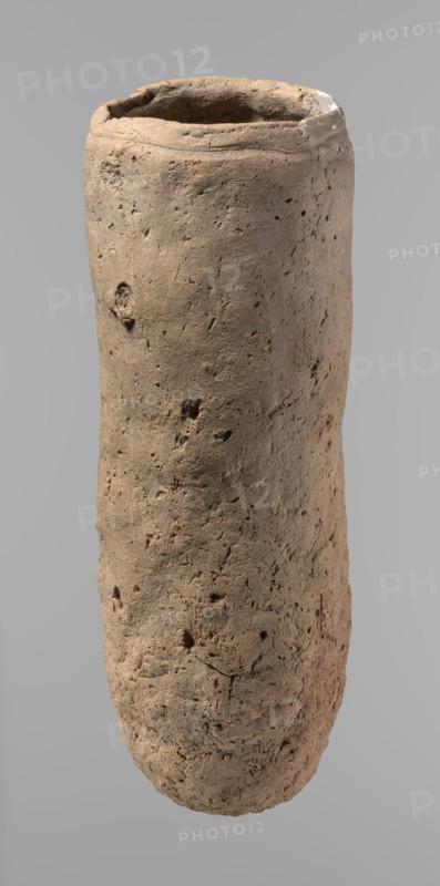 Vessel, Egyptian pottery, handmade clay vessel, almost cylindrical, widest just below the rim, rounded bottom, slightly marked narrow sloping shoulders, straight tapering rim. Handmade of Nile clay with numerous organic inclusions and limestone particles visible on the surface; surface uneven and better worked in the upper part, with numerous uneven depressions especially at the bottom. Color brown. Fairly well preserved; cracks on the surface attributed to careless craftsmanship during kneading and firing. Old Kingdom, ca. 2686–2181 BC; Edfu place of origin. Vessel; height 36.5 cm, diameter 11.5 cm rim, diameter 13.2 cm body., A tall cylindrical object with a rough, pitted surface in shades of beige and light brown, showing irregular dents, holes, and small cracks; the top edge is uneven and slightly thicker with a darker interior, and the background is a smooth neutral gray.