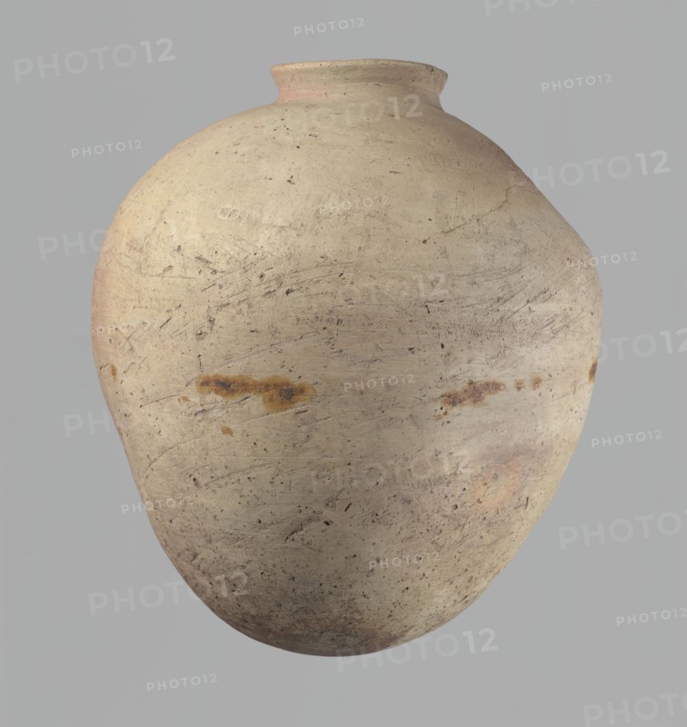 Vessel, Vessel of an ovoid, slightly downward-stretching shape, widest at about 2/3 of the height. Broad, sloping shoulders, very short, wide neck. Rounded rim, slightly profiled in the lower part, oval in cross-section, slightly flaring. Rounded bottom. Just above the widest diameter of the vessel there is a dent made before the vessel was fired. Clay NB2 according to the Vienna System with a significant amount of organic admixture. The vessel was wheel-made, with approximately half of its height worked with a knife or similar tool. The vessel is beige. The vessel is cracked in the upper part. c. 2686–c. 2181 BC; Old Kingdom, Edfu; vessel. Height 34 cm; rim diameter 10.5 cm; body diameter 29 cm., A large rounded pottery vessel with a narrow short neck and flared rim sits against a plain background; the vessel surface is a weathered beige with areas of light tan and subtle pinkish hues, and there are a few rusty brown stains and darker speckles scattered across its textured, slightly s...