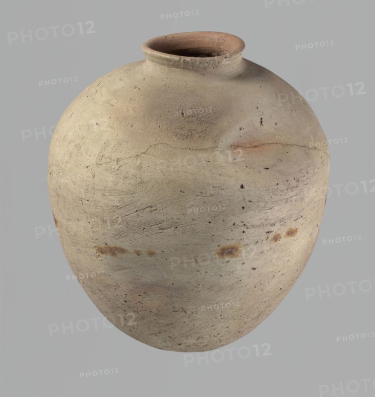 Vessel, Egyptian pottery, ceramics, clay vessel. Ovoid, slightly downward-stretching shape, widest at about two-thirds of the height; broad, sloping shoulders; very short, wide neck; rounded rim, slightly profiled in the lower part, oval in cross-section, slightly flaring; rounded bottom. Dent just above the widest diameter made before firing. Clay NB2 Vienna System with significant organic admixture. Wheel-made; approximately half the height worked with a knife or similar tool. Beige. Cracked in the upper part. Old Kingdom, c. 2686–c. 2181 BC; from Edfu. Vessel; height 34 cm; rim diameter 10.5 cm; body diameter 29 cm., A rounded clay vessel with a narrow neck and flared rim, showing a textured, uneven surface with a few rough patches and small pits; the body displays subtle horizontal tool marks and faint surface cracks, along with several darker spots and smudges. The visible colors include light beige, pale tan, muted brown, touches of warm reddish-brown, and small areas of darker b...