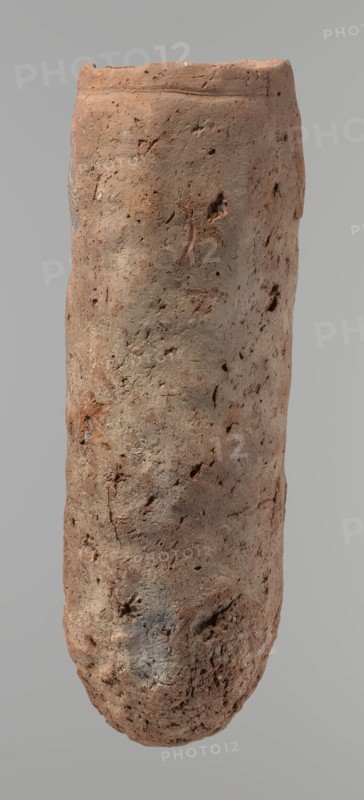 Vessel, An almost cylindrical handmade Nile-clay vessel, widest just below a straight, slightly tapering rim; rounded bottom; shoulders almost illegible. Made of not-well-worked Nile clay with numerous organic inclusions and limestone particles visible on the surface. Carelessly made with uneven depressions, especially at the bottom; brown in color; cracks on the surface due to workmanship and firing. Ceramic vessel, clay; Old Kingdom, ca. 2686–ca. 2181 BC; Edfu. Height 37.9 cm; diameter 11.4 cm rim, 12.5 cm body., An elongated cylindrical clay object with an uneven, rough surface showing a mix of reddish-brown, tan, gray, and faint darker brown tones, pitted and marked with small holes, cracks, and irregular indentations across its surface, with a slightly flattened top rim and a rounded bottom, photographed against a plain light gray background.