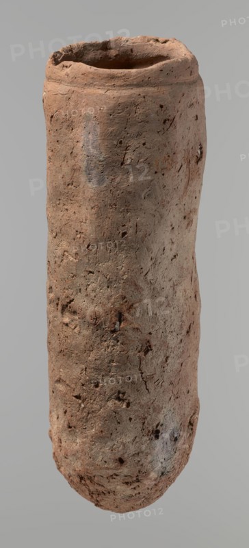 Vessel, Egyptian pottery, handmade clay vessel, almost cylindrical, widest just below a straight, slightly tapering rim; rounded bottom; shoulders almost illegible. Handmade from Nile clay with numerous organic inclusions and limestone particles visible on the surface; carelessly made with uneven depressions especially at the bottom; brown color; cracks from clay preparation and firing. Date ca. 2686–ca. 2181 BC Old Kingdom, from Edfu. Vessel, height 37.9 cm; diameter 11.4 cm rim, 12.5 cm body., A tall cylindrical clay vessel with an irregular rim and rough, pitted surface, showing a weathered texture and small holes and indentations across its body; the form is slightly tapered with subtle bulges and a rounded base, and visible marks and scratches on the exterior. The colors visible are various shades of brown, reddish-brown, gray, and a few darker brown spots, set against a neutral light gray background.