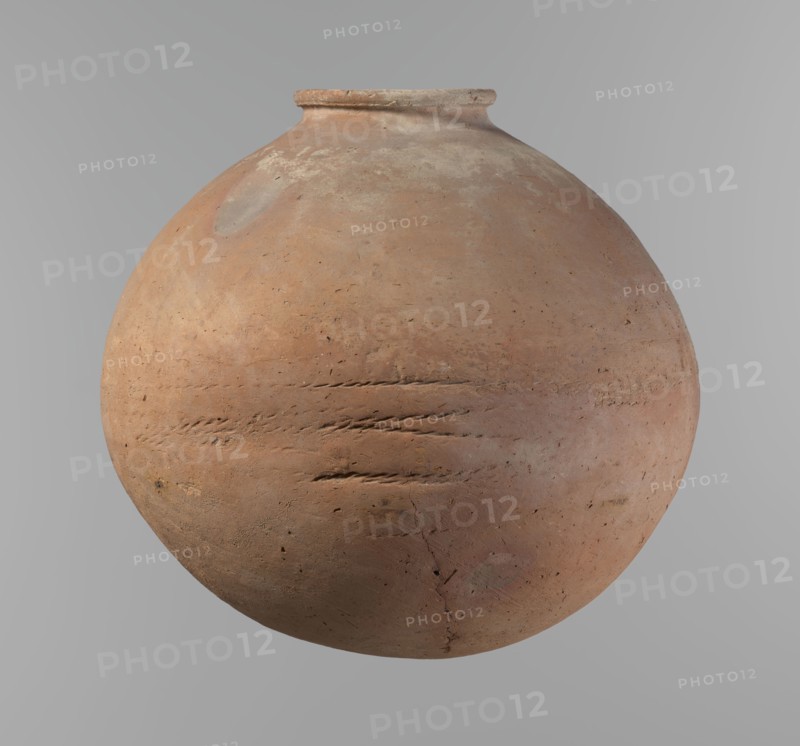 Vessel, Egyptian pottery vessel with an almost spherical, slightly horizontally stretched shape, widest about half the height. Broad, sloping shoulders, very short neck, rounded rim oval in cross-section, slightly flaring and rounded bottom. Horizontal cord impressions at the widest point. NB2 clay Vienna System with a significant amount of organic admixture. Surface smooth. Made on a wheel; approximately half of its height was worked with a knife or similar tool. Beige color. Very well preserved. Date: ca. 2686–ca. 2181 BC Old Kingdom, from Edfu. Height 28.5 cm; rim diameter 10.4 cm; body diameter 30.410 cm., A large round clay vessel with a narrow rim floating against a plain light gray background, showing a surface of earthy terracotta with areas of pale beige and muted pinkish tones, subtle gray patches, and darker brown spots; the vessel bears rough textured marks, scratch-like grooves, small pits, and a visible crack, with overall matte, weathered appearance and uneven coloration...