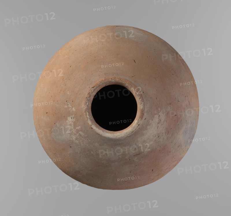 Vessel, Egyptian pottery ceramic clay vessel. A vessel with an almost spherical, slightly horizontally stretched shape, widest about half the height. Broad, sloping shoulders, very short neck. Rounded rim, oval in cross-section, slightly flaring. Rounded bottom. Horizontal cord impressions at the widest point of the vessel. NB2 clay according to the Vienna System with a significant amount of organic admixture. The surface is smooth. The vessel was made on a wheel, and approximately half of its height was worked with a knife or similar tool. Colour beige. Very well preserved. Ca. 2686–2181 BC; Old Kingdom, Edfu. Vessel height 28.5 cm; rim diameter 10.4 cm; body diameter 30.410 cm., A round, top-down view of a large clay vessel with a central circular opening, showing a rough, textured surface in shades of brown, tan, reddish-brown, gray, and muted beige, with a few darker spots and subtle lighter patches, set against a smooth light-gray background.