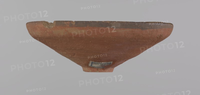 Bowl, Egyptian painted pottery bowl with conical shape and ring-shaped foot. Straight rim, slightly flaring outward, irregular. Wheel-made of Nile clay type ND Vienna System. Surface even, covered with glaze before firing; inner and outer rim bear a narrow ˜0.5 cm black band; remainder of vessel is red. Traces of polish; glaze slightly worn in places, possibly from use. Good condition with minor clay chips on rim and abrasions over surface. Analogies: Egypt's Golden Age no. 51, p. 78, ca. 1550–ca. 1425 BC; New Kingdom, early 18th Dynasty. Provenance notes: Edfu, Deir el-Medina . Vessel. Height 5.3 cm; rim diameter 15.1 cm; bottom diameter 3.1 cm., A shallow, wide bowl viewed from the side showing its rounded exterior and narrow base, with a rough, weathered surface and a chipped rim; the bowl is primarily reddish-brown with darker brown along the rim and a few lighter tan patches, set against a pale gray background.