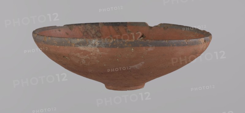 Bowl, A conical clay bowl with a ring-shaped ring bottom, wheel-made from Nile clay type NB2, Vienna System. The walls are even and covered with red engobe; the inner surface is polished. The rim is straight, slightly curled toward the center. A black painted band approx. 1 cm wide appears on the inside and outside of the rim. Vessel color: red. Preservation: quite good, minor losses on the rim and minor surface damages. Comparative dating: New Kingdom, 18th Dynasty ca. 1550–1295 BC; Tuthmosis III. Dimensions: height 5.5 cm; rim diameter 16.4 cm; bottom diameter 3.6 cm. Place of origin: Deir el-Medina, Egypt., A shallow, rounded ceramic bowl with a worn rim and uneven edge, showing surface chips and weathering; the bowl's exterior and interior display a terracotta color with patches of darker brown and areas of lighter, pale discoloration; the rim has a band of dark grayish-brown with small flecks of yellowish residue; the surface texture appears rough and pitted in places, with a smal...