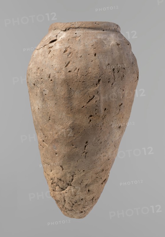 Vessel, Egyptian pottery, hand-made clay vessel, Old Kingdom c. 2686–c. 2181 BC, Edfu, vessel height 31.9 cm, rim diameter 10.9 cm, body diameter 17.4 cm., A single large clay vessel with a rough, pitted surface occupies the image, shown against a plain light gray background; the vessel has an elongated, tapered shape with a rounded shoulder and a slightly flaring rim, and its surface shows many small holes, cracks, and irregular textures. The visible colors are various shades of brown on the vessel, with subtle spots of darker brown and hints of gray, and the background is a uniform light gray.