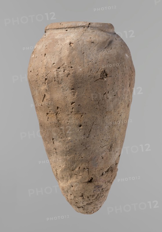 Vessel, Egyptian pottery, hand-made clay vessel; Old Kingdom, c. 2686–c. 2181 BC; from Edfu; vessel height 31.9 cm, rim diameter 10.9 cm, body diameter 17.4 cm. Comparative material: Petrie, Dendera, pl. XVI,2; Hope, Akhmim, fig. 7, type AIVa 2bi; Reisner, Mycerinus, p. 212, fig. 64 3,4; Brunton, Qau and Badari, pl. LXXXI type 77N — earlier dated, to the IVth–Vth Dynasty; Mace, Naga ed-Der II, figs. 87.11–14, 16–25; Reisner, Naga ed-Der III, fig. 28–33; Petrie, Abydos II, pl. XLIII, 79–80; Keiser, Avila, Dreyer, Jaritz, Laskowska-Kusztal, MDAIK 40, fig. 9b, c., A large, weathered clay vessel with a rounded shoulder tapering to a pointed base, shown against a plain light gray background. The surface of the vessel is rough and pitted with irregular indentations, small holes, and fine scratches. The color palette is mainly earthy tones including light brown, tan, beige, and hints of darker brown in textured areas. The rim is slightly uneven and shows wear and small chips. There are areas ...