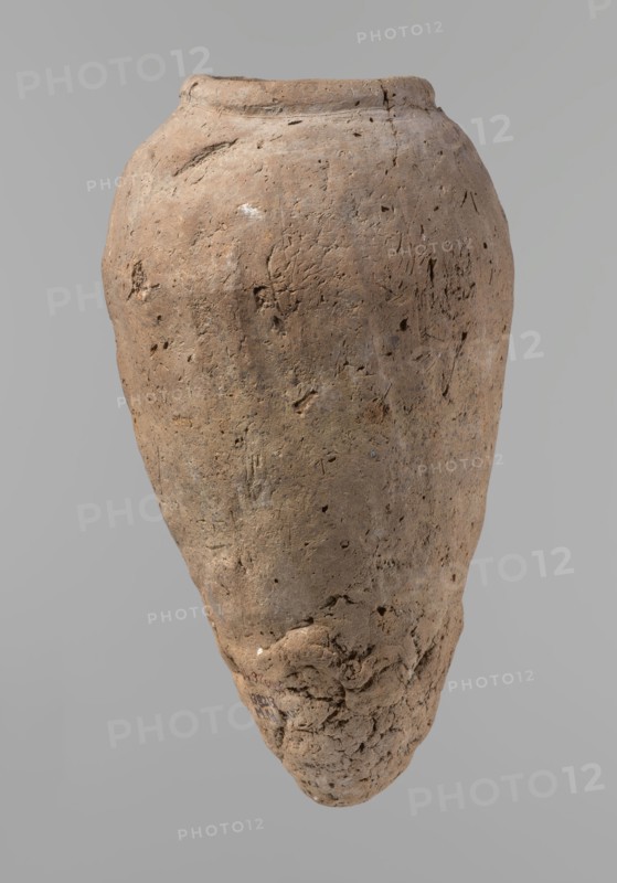 Vessel, Egyptian pottery, hand-made clay vessel, ceramics; comparative material: Petrie, Dendera, pl. XVI,2; Hope, Akhmim, fig. 7, type AIVa 2bi; Reisner, Mycerinus, p. 212, fig. 64 3,4; Brunton, Qau and Badari, pl. LXXXI type 77N - earlier dated to the IVth–Vth Dynasty; Mace, Naga ed-Der II, figs. 87.11–14, 16–25; Reisner, Naga ed-Der III, fig. 28–33; Petrie, Abydos II, pl. XLIII, 79–80; Keiser, Avila, Dreyer, Jaritz, Laskowska-Kusztal, MDAIK 40, fig. 9b,c. c. 2686–c. 2181 BC; Old Kingdom, Edfu; vessel height 31.9 cm, rim diameter 10.9 cm, body diameter 17.4 cm., A large, weathered clay vessel with an uneven, rounded body and a narrow, rough base is shown against a plain light gray background; the surface of the vessel is textured and pitted with irregular indentations and small holes, and it displays a range of earthy browns with subtle patches of darker and lighter brown, a few pale beige and off-white spots, and faint areas of grayish discoloration.
