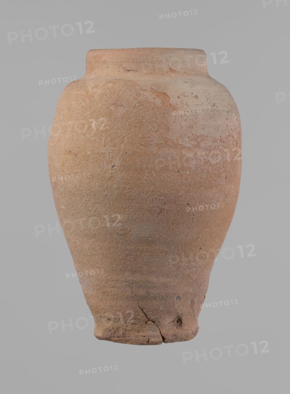 Vessel, Beer jug of a slightly irregular, conical shape with a wide, very short neck and a flat bottom. Flat, narrow shoulders and a gently rounded, straight rim. Wheel-made of Nile clay type NB2 Vienna System. Bottom cut off with string and bearing clear potter's fingerprints, flaring slightly in its lowest part. Surface even. Color: red. Complete and fairly well preserved, with minor damage to the bottom. New Kingdom, ca. 1550–1069 BC, provenance: Edfu. Dimensions: height 24 cm; rim diameter 10 cm; body diameter 16 cm; bottom diameter 9.2 cm., A large rounded ceramic vessel with a narrow flat rim and a tapered base sits against a plain background; its surface shows a rough, grainy texture with areas of unevenness and small pits, and there are visible cracks and chips near the base and along the body. The vessel displays earthy tones of brown, tan, beige, and hints of reddish-orange, with subtle variations and mottling across the surface.