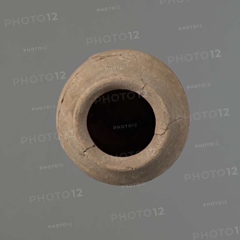 Vessel with hieratic inscription, Egyptian pottery, ceramics, clay conical vessel tapering towards the bottom, irregular, widest approx. 3/4 of the height. Narrow, slightly sloping shoulders; short, wide neck; very gently rounded, slightly flaring rim; pointed bottom. Shallow horizontal lines at the widest diameter made before firing. Handmade vessel; upper part shoulders, neck, rim turned. Even surface, outer probably covered with a layer of engobe. Made of Nile clay NB2 Vienna System. Remains of a hieratic inscription in black on lower part of the belly. The vessel is glued from original fragments. Old Kingdom, Edfu. Height 14.8 cm; rim diameter 6.2 cm; belly diameter 9.1 cm. Comparable types dated ca. 2686–ca. 2181 BC; earlier parallels assigned to 4th–5th Dynasty., A round, weathered clay vessel viewed from above with a wide dark opening at its center, showing a rough, cracked surface and uneven rim; the vessel is primarily brown with variations of tan and gray, and it sits against...