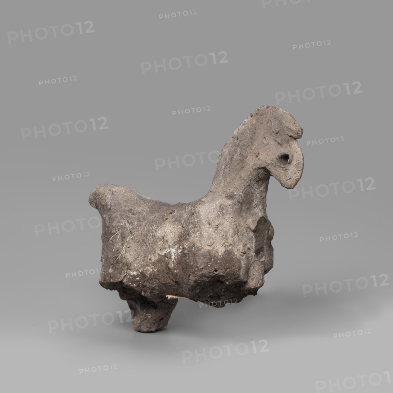 Horse figurine. Schematic representation of a horse with a short torso, a massive arched neck and a crest-like mane; small head with eyes marked by a drilled hole; raised tail preserved in the upper part. Late Period–Ptolemaic Period, 664–30 BC. From Edfu. Terracotta figurine, height 8 cm, width 9 cm., A small sculptural object resembling an animal form sits against a smooth gray background; the piece appears rough and irregular with a textured surface showing wear and pitting, and its overall color palette includes shades of gray, brown, and muted beige. The object has a raised head with a rounded snout and a hollowed area where an eye might be, a short neck with a scalloped ridge along the top, a broad body that tapers toward a stubby tail, and short, thick legs providing support. Surface variations show lighter and darker patches, small pits, and areas of rougher, crumbly texture, with some crusty deposits and subtle scratches visible on the body.