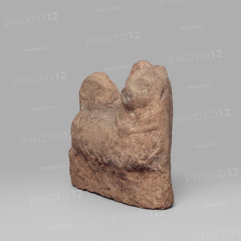 Dog figurine, Roman period, 1st–2nd century; found at Edfu; height 6.6 cm, width 6.5 cm., A roughly triangular, weathered brown clay object with an uneven, textured surface stands upright against a plain gray background, showing a few rounded protrusions at the top and a rough, irregular base with varied brown and reddish-brown tones.