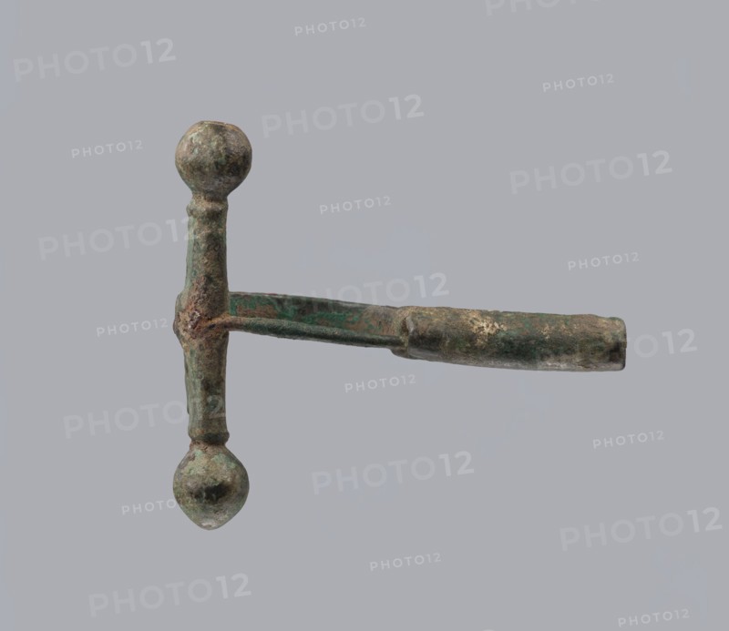 Onion-shaped fibula, Italy. Bronze fibula with hinged structure: an arched ring made from a quadrilateral plate, one end terminating in a thick transverse plate with a stepped upper edge and large balls on both sides, the other end a thick sheath shaped as a half-cylinder, cut to insert a needle. Condition: good; surface covered with greenish corrosion and brown spots. Late 3rd–4th century. Jewelry, fibula., An object with a vertical shaft and a horizontal arm forming a T-shape, showing rounded knobs at the top and bottom of the shaft and a tubular extension on the horizontal arm; the surface appears rough and corroded with mottled green and brown patina and some darker and lighter patches, set against a uniform light gray background.