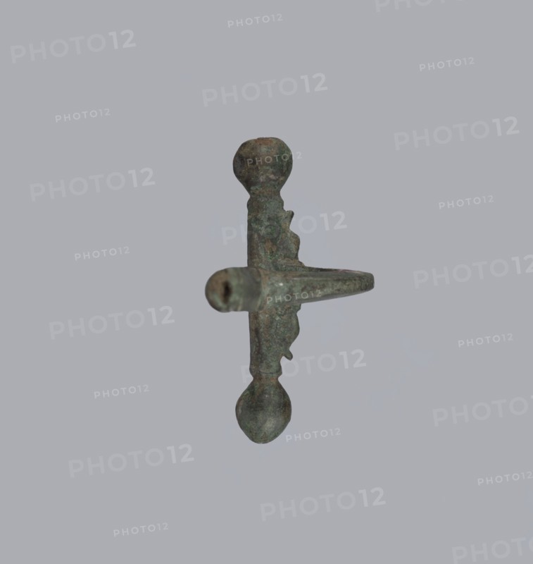 Onion-shaped fibula, Italy; bronze fibula with hinged structure: an arched ring formed from a quadrilateral plate, one end terminating in a thick transverse plate with a stepped upper edge and large balls on both sides, the other ending in a thick sheath shaped as a half-cylinder, cut to receive a needle. Condition: good, surface with greenish corrosion and brown spots. Date: late 3rd–4th century; probable origin: Rome. Jewelry, fibula., A single metal object is centered against a plain light gray background, showing a vertical shaft with rounded knobs at both ends and a horizontal protruding arm; the object has a weathered, mottled surface with greenish patina and areas of brown and dark gray, and irregular textured edges and pitted spots are visible along its form.