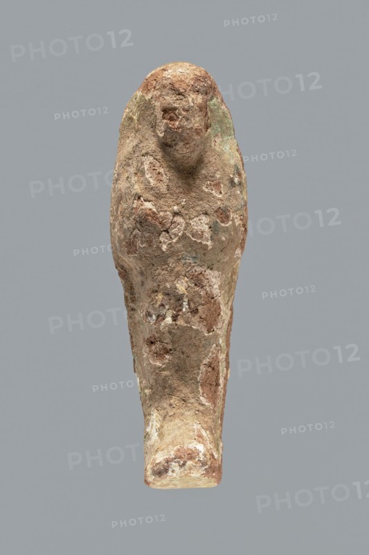 Ushabti. A mummy-shaped ushabti figurine made of clay, impressed in a mold. Traces of white primer and paint in places on the surface. The back of the figurine is unfinished. The arms are separated, the hands are crossed on the chest, probably the left folded over the right. In both hands the figurine holds hoes — the right much more clearly visible, the left very indistinct. The tools are painted black. A three-part wig on the head, the strand at the back is not separated. The ears are invisible. Facial details illegible. Inscription illegible/missing. Figurine not very well preserved — significant paint loss over the entire surface. Typology according to Schneider: 4.3.2 VIIIA2/W.16 H.29 I.5 B.0 Tp.0. ca. 1069–715 BC; Third Intermediate Period, 21st–23rd Dynasty ca. 1069–715 BC, Egypt. Shabti. Height 9.4 cm; width 3.1 cm; depth 2.7 cm., A weathered vertical sculpture of a simplified human figure with a rounded head and indistinct facial features, displaying a rough, pitted surface an...