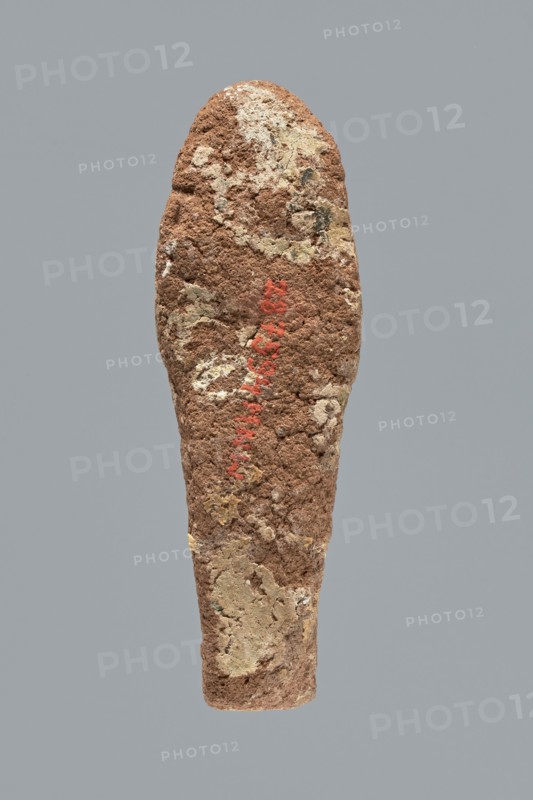 Ushabti. A mummy-shaped ushabti figurine made of clay, impressed in a mold. Traces of white primer and paint remain in places; significant overall paint loss. The back is unfinished. Arms are separate, hands crossed on the chest with the left likely folded over the right. The figurine holds hoes in both hands — the right hoe is much clearer, the left indistinct; the tools are painted black. Head bears a three-part wig with the rear strand not separated; ears invisible; facial details illegible. Inscription illegible/missing. Typology Schneider: 4.3.2 VIIIA2/W.16 H.29 I.5 B.0 Tp.0. Date ca. 1069–715 BC, Third Intermediate Period 21st–23rd Dynasty. Type: shabti. Dimensions: height 9.4 cm, width 3.1 cm, depth 2.7 cm., A roughly elongated, tapering object with a textured, crumbly surface in shades of brown and tan, with patches of lighter beige and pale yellow; there is a vertical area of muted red pigment on the surface; the background is a flat, uniform gray.
