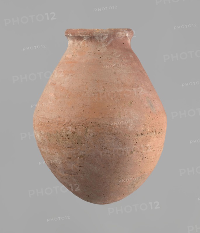 Vessel, Egyptian ceramics. Wheel-made from Nile clay of type NB2 Vienna System. Ovoid belly, wide short neck, rounded bottom, straight upturned rim. Surface levelled and covered with engobe; outer surface red, inner surface yellowish-brown. Firing hole at widest circumference, approx. 1 cm diameter. Well-preserved. ca. 1550–1295 BC; New Kingdom, 18th Dynasty; Deir el-Medina. Vessel: height 27 cm; rim diameter 11 cm; body diameter 20.5 cm., A large rounded clay vessel with a narrow neck and slightly flared rim is centered against a plain light gray background; the surface of the vessel shows a rough, textured finish with visible small pits and irregularities and a gradient of earthy colors including terracotta, muted orange, pale pink, and areas of dusty brown and gray, with subtle darker speckles and weathered patches.